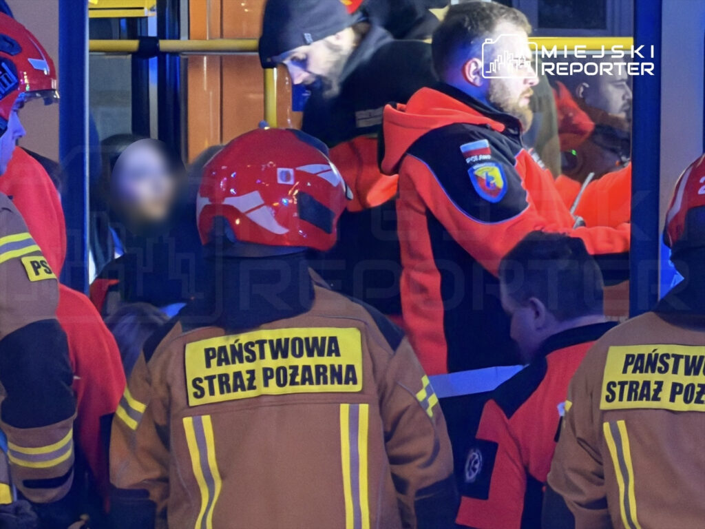 Grupa ratowników w hełmach udziela pomocy osobie w pojeździe w nocy, otoczeni przez innych pracowników służb.