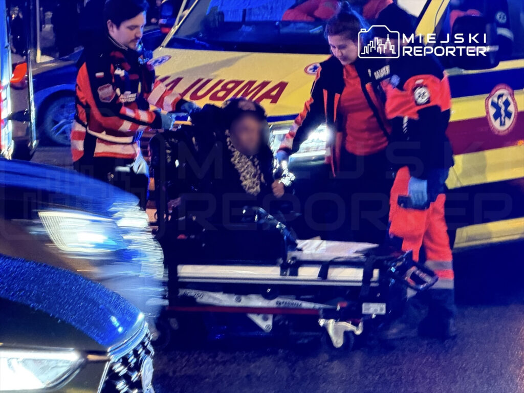 Ratownicy medyczni udzielają pomocy osobie na wózku inwalidzkim obok karetki w nocy, w ruchliwym miejscu.