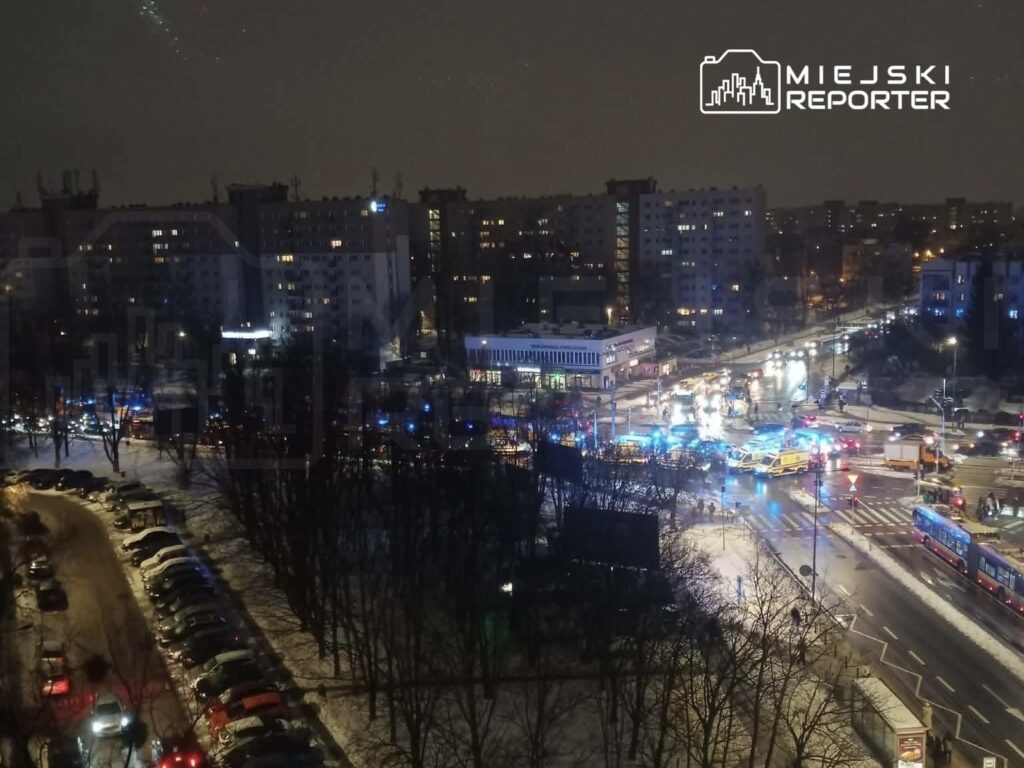 W nocy, na ruchliwym skrzyżowaniu, widoczne są pojazdy służb ratunkowych oraz oświetlenie w kolorze niebieskim i czerwonym.