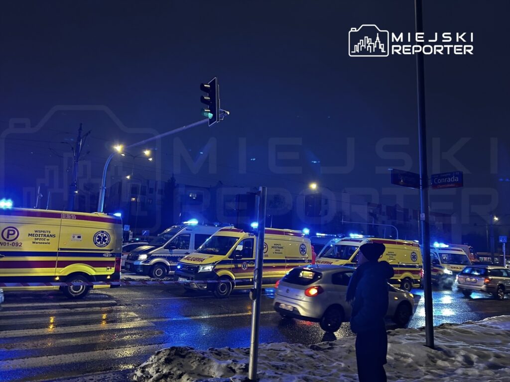 W nocy na skrzyżowaniu z sygnalizacją świetlną stoją karetki pogotowia, a przechodzień obserwuje sytuację.