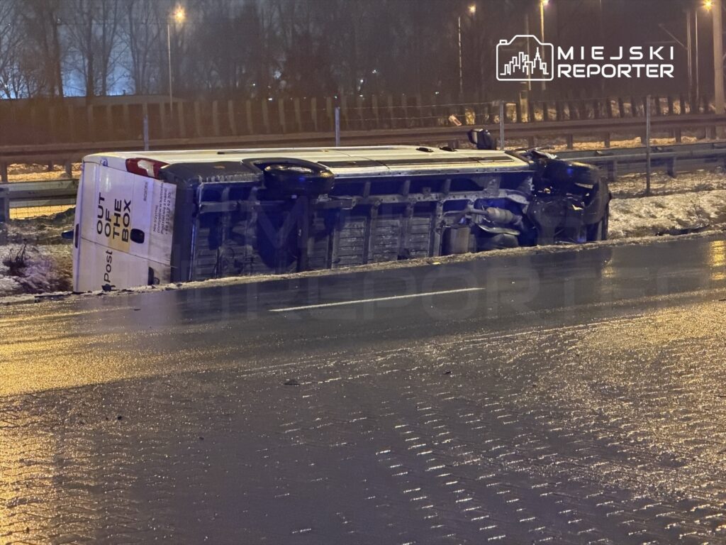 Przewrócony samochód dostawczy leży na boku na poboczu drogi, obok widoczne są zmarznięte trawy i latarnie uliczne.