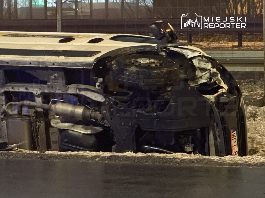 Przewrócone auto osobowe leży na boku na poboczu drogi, obok widoczny jest zarys asfaltu i śniegu.