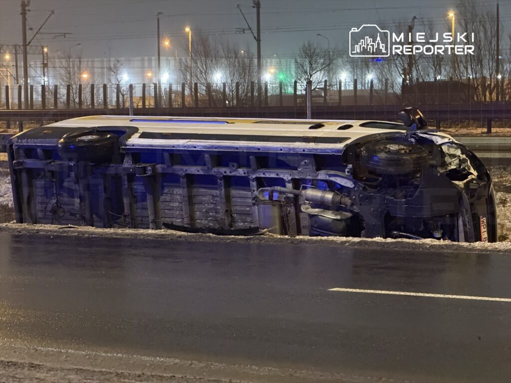 Przewrócony autobus leży na boku na drodze, w tle widać oświetlone drzewa i infrastrukturę miejską.