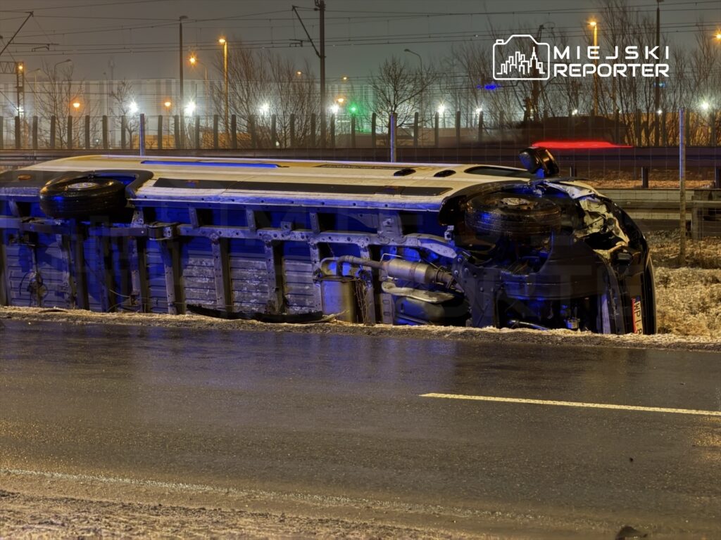 Przewrócony pojazd leży na boku na drodze, otoczony przez nocne oświetlenie i zaspy śnieżne.