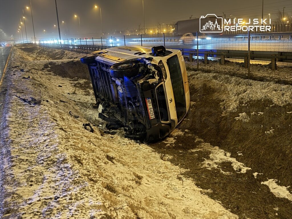 Przewrócone auto osobowe leży na poboczu drogi, obok widoczne fragmenty zderzenia, nocna sceneria z oświetleniem ulicznym.