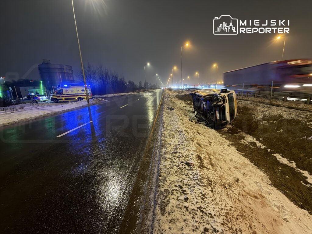 Na drodze leży przewrócony samochód, obok widać radiowóz i karetkę z włączonymi sygnałami świetlnymi.