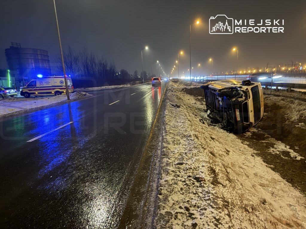 Na poboczu drogi leży przewrócony samochód, obok stoi karetka pogotowia, a w tle widoczne są światła innych pojazdów.