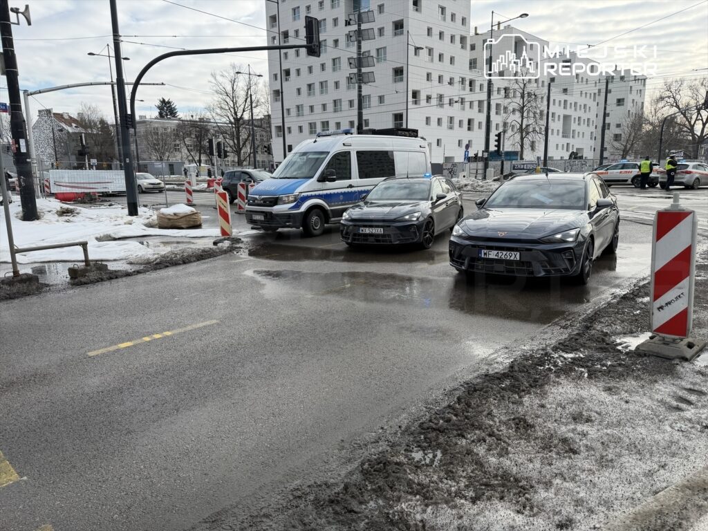 Policyjny van oraz dwa czarne samochody osobowe stoją na drodze, w tle widać nowoczesne budynki i plac budowy.
