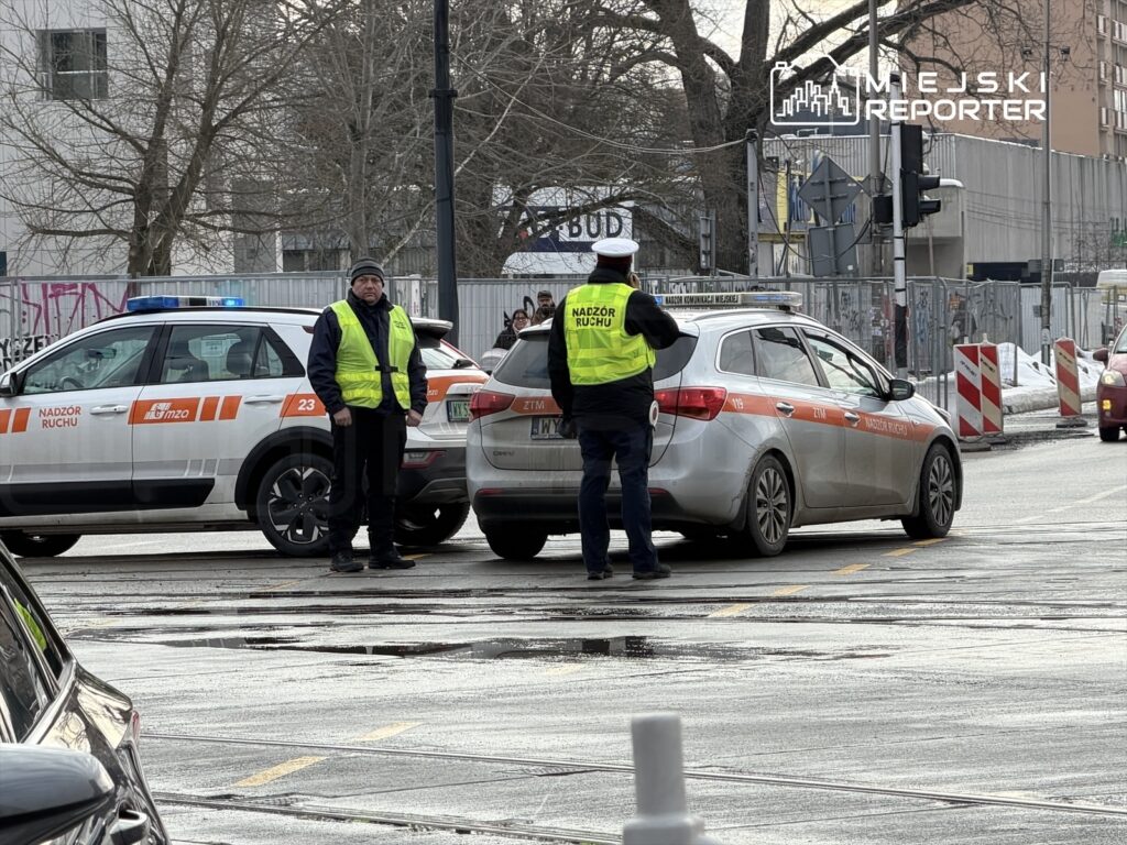 Dwaj mężczyźni w kamizelkach odblaskowych kontrolują ruch przy skrzyżowaniu, obok stoi samochód z napisem "Nadzór Ruchu".