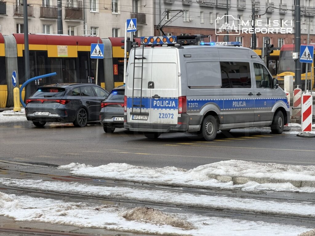 Radiowóz policyjny z włączonymi sygnałami świetlnymi stoi obok samochodu osobowego na zablokowanej ulicy.