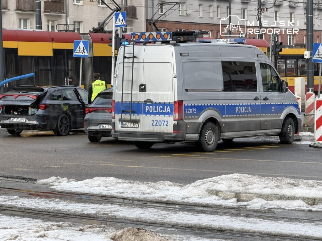 Policjant w żółtej kamizelce rozmawia z kierowcą przy czarnym samochodzie osobowym obok radiowozu.