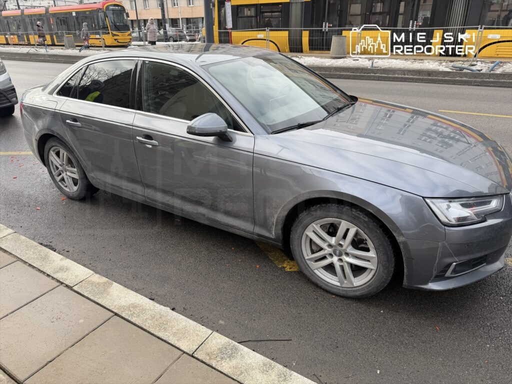 Szary samochód osobowy zaparkowany na ulicy, obok widoczny tramwaj w ruchu oraz piesi na chodniku.