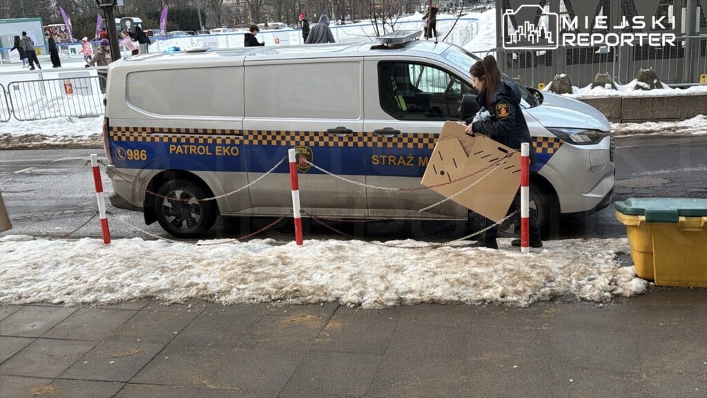 Funkcjonariuszka straży miejskiej trzyma karton na tle radiowozu z napisem "PATROL EKO" obok śnieżnego chodnika.