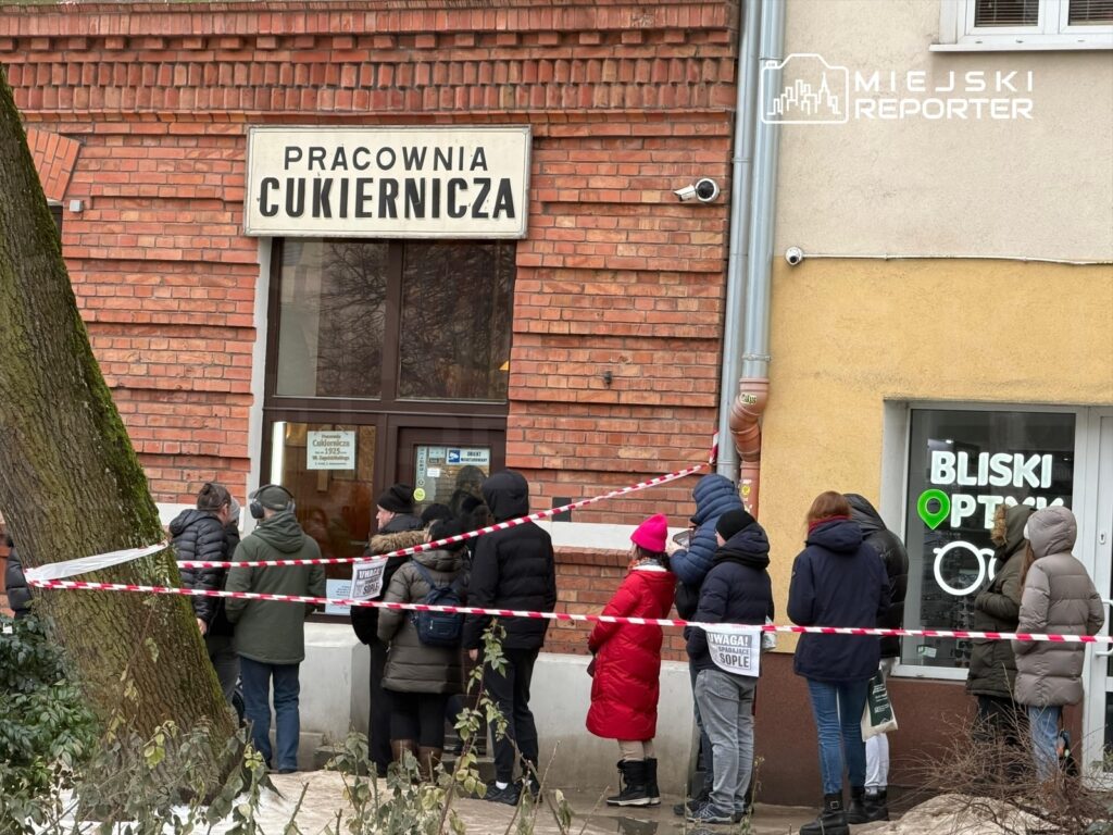 Ludzie stoją w kolejce przed cukiernią, obok widoczna jest żółta ściana z oknem optyka.