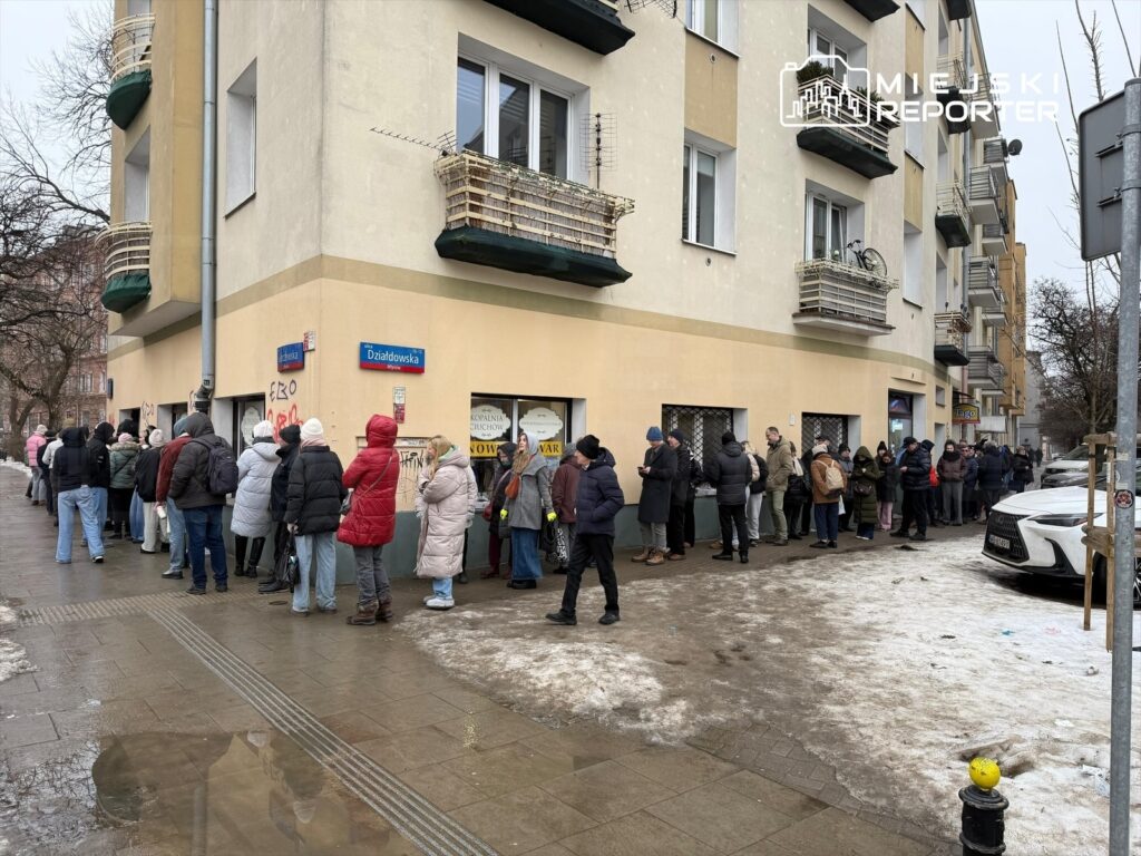 Ludzie w różnych kurtkach stoją w długiej kolejce przed budynkiem z oknami zabezpieczonymi kratami.