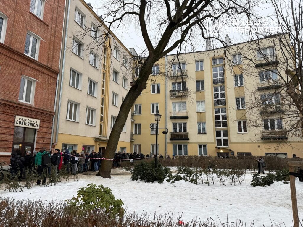 Ludzie stoją w kolejce przed cukiernią, obok nowoczesnych budynków, na śnieżnym podłożu.