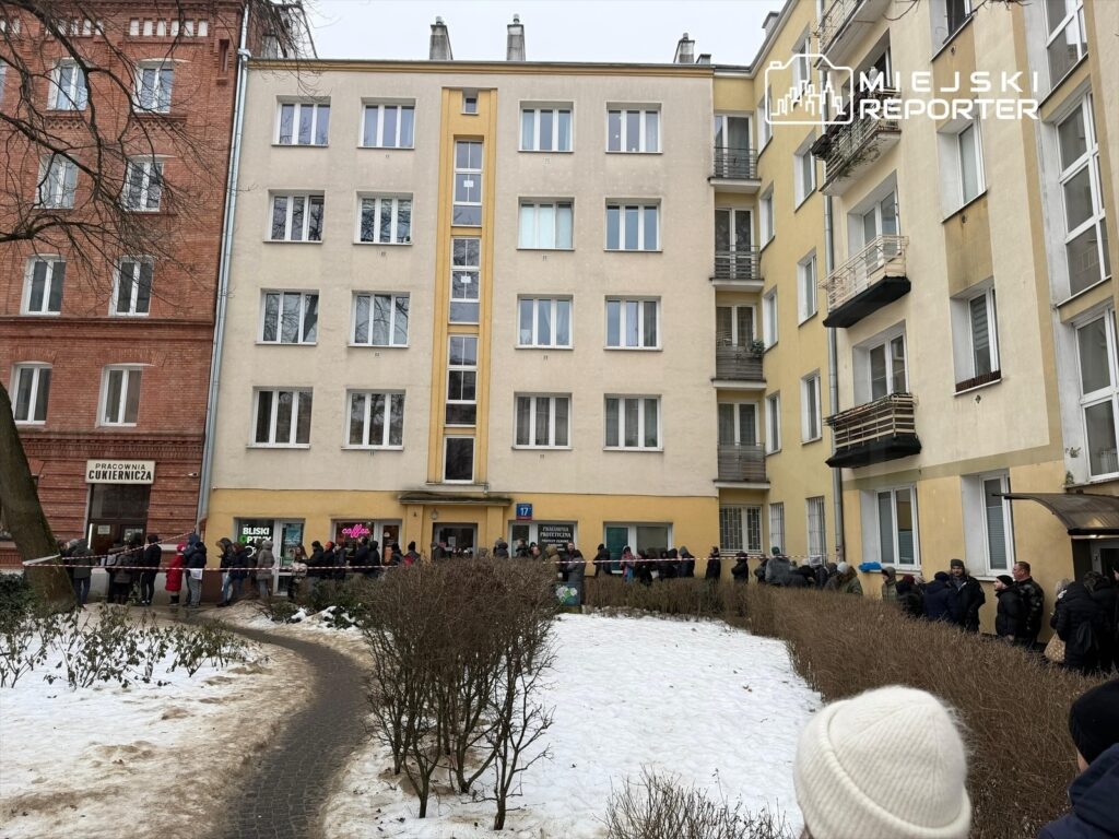 Ludzie stoją w długiej kolejce przed budynkiem mieszkalnym w zimowej scenerii, otoczeni śniegiem i krzewami.