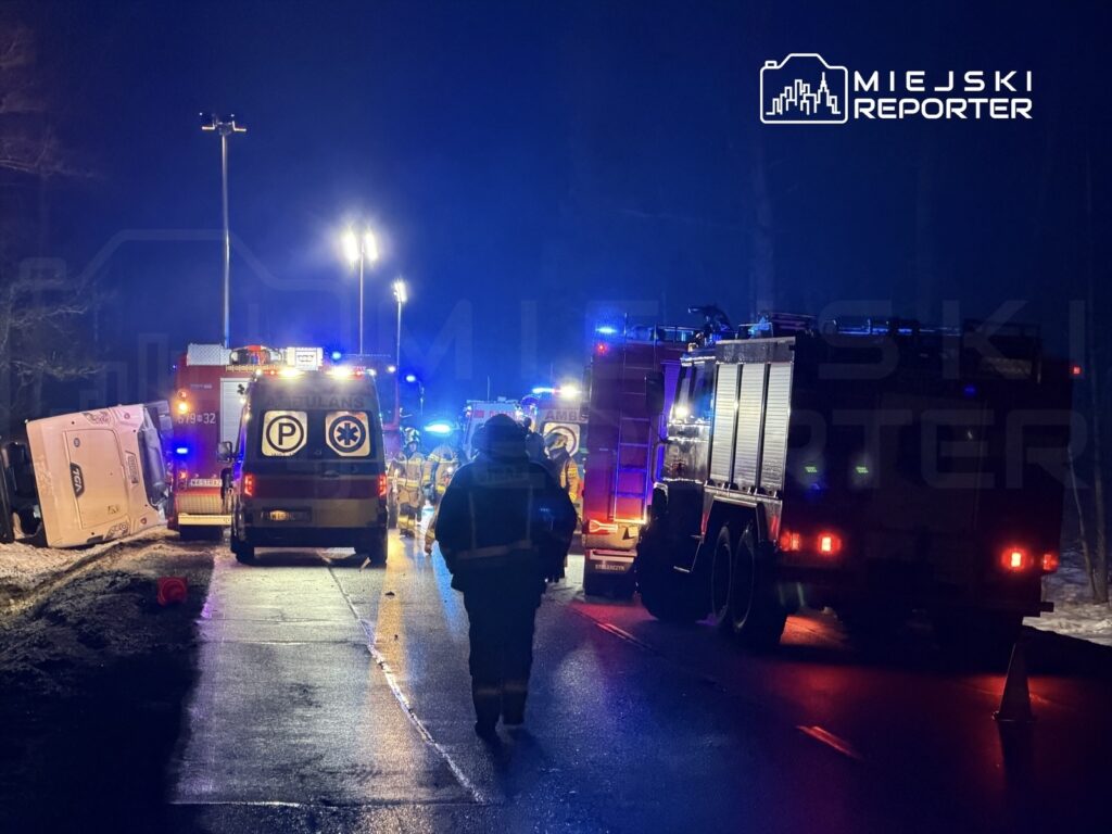 Na drodze nocą, służby ratunkowe pracują przy przewróconym pojeździe; w tle widać wozu strażackie oraz karetki.