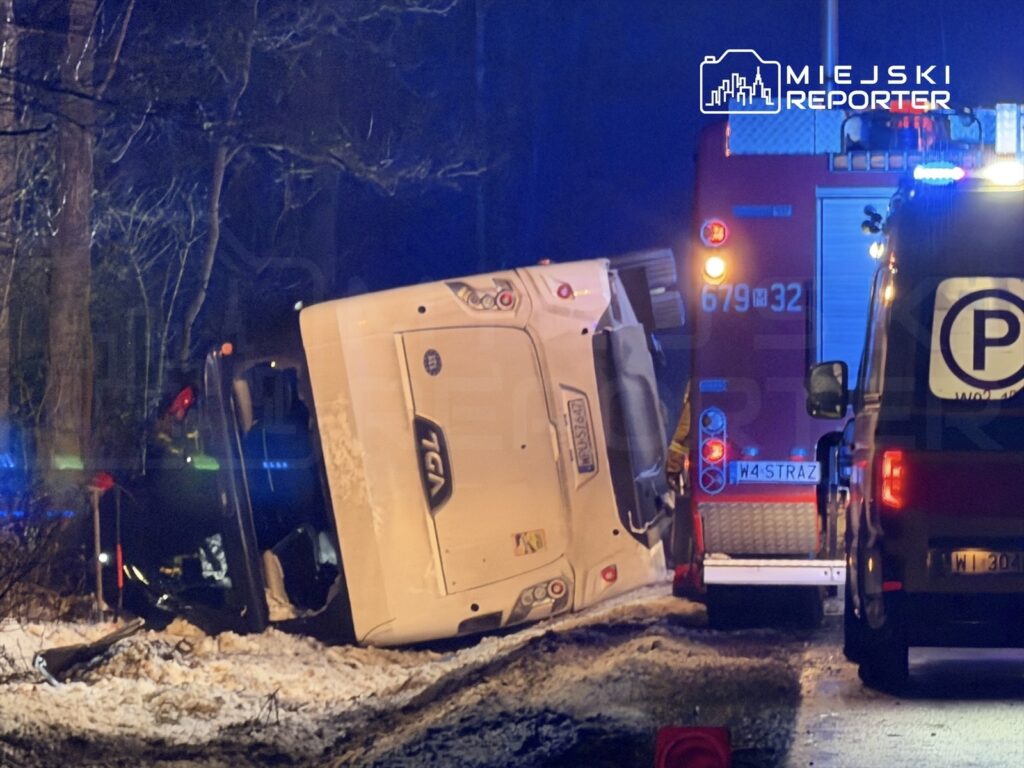Uszkodzony pojazd leży na boku przy drodze w lesie, obok stoją pojazdy straży pożarnej z włączonymi sygnałami.