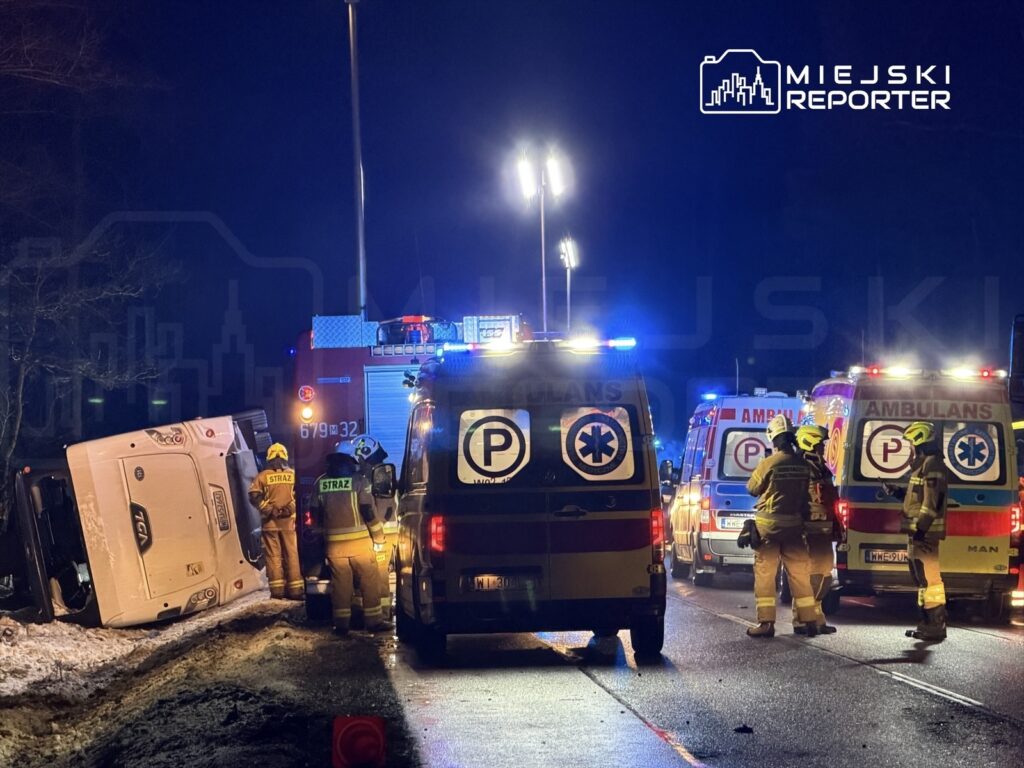 Strażacy i ratownicy medyczni pracują przy przewróconym pojeździe na poboczu drogi w nocy, otoczeni przez pojazdy ratunkowe.
