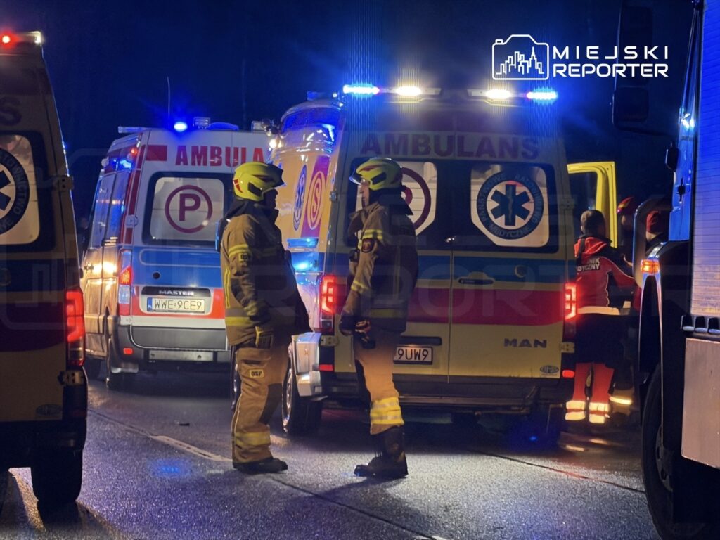 Ratownicy medyczni w hełmach rozmawiają obok ambulansów z włączonymi sygnalizatorami na nocnej drodze.