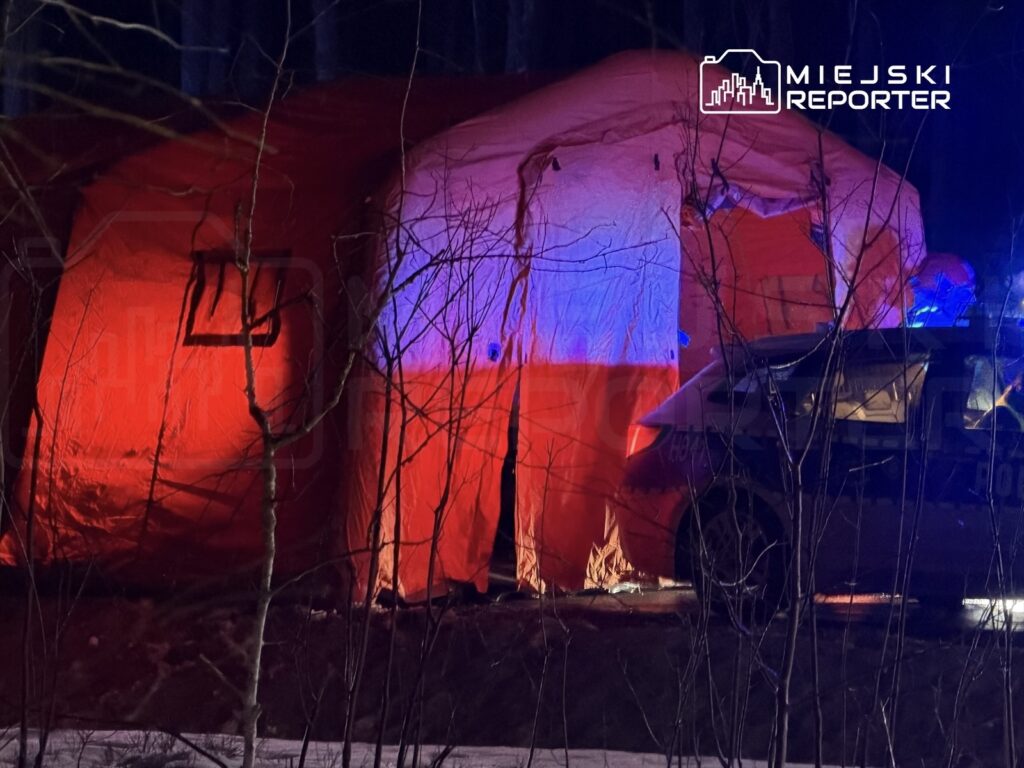 Czerwony namiot ratunkowy oświetlony niebieskimi i czerwonymi światłami, obok policyjny samochód w lesie.