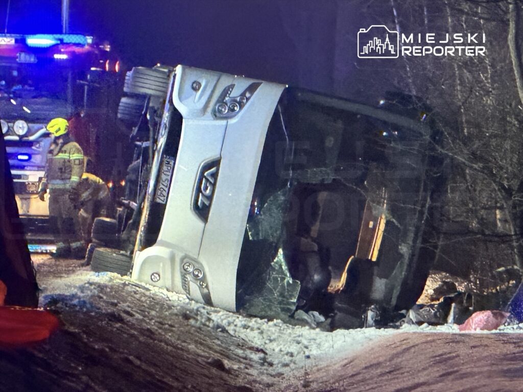 Wydobyty pojazd osobowy leży na boku na zaśnieżonej drodze, w tle strażacy oraz pojazd ratunkowy z włączonymi sygnałami świetlnymi.