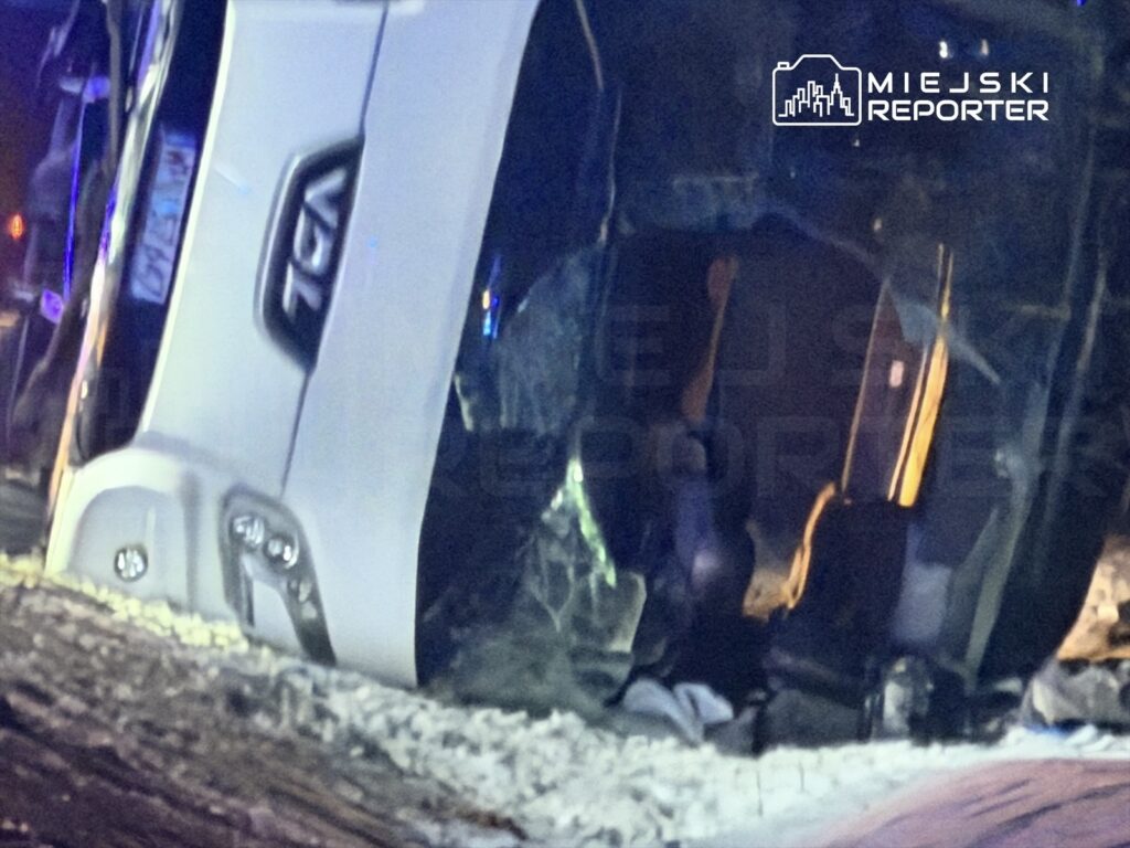 Przewrócony samochód osobowy leży na boku na śniegu, w tle widać migające światła policyjne.