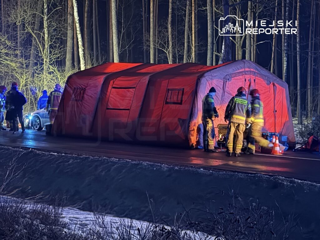Grupa ratowników w odblaskowych strojach pracuje przy czerwonym namiocie na drodze, otoczeni przez drzewa w nocy.