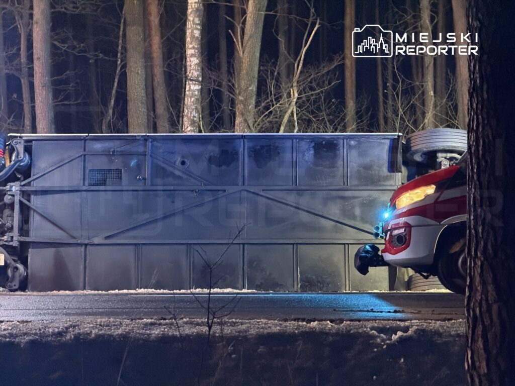 Na drodze leży przewrócony pojazd, a obok niego znajduje się samochód ratunkowy z włączonymi światłami. W tle widoczne są drzewa.