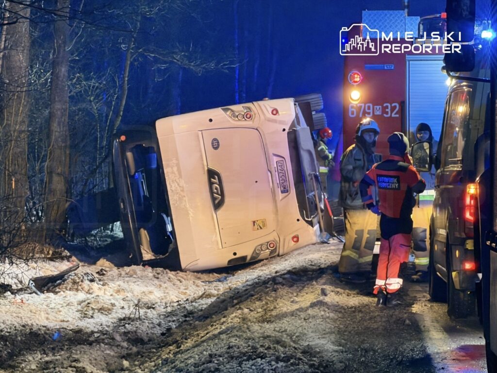 Wielki autobus leży na boku w lesie, obok stoją ratownicy medyczni i strażacy, w tle widać pojazdy ratunkowe.
