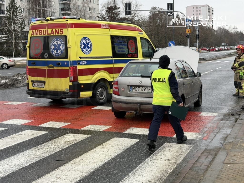 Ambulans z włączonymi sygnałami świetlnymi stoi na drodze, obok samochód z otwartą maską; policjant w kamizelce odblaskowej prowadzi działania.