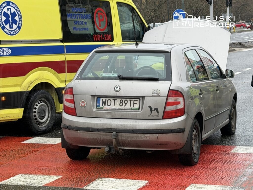 Srebrny samochód osobowy z otwartą maską stoi na przejściu dla pieszych obok karetki pogotowia.