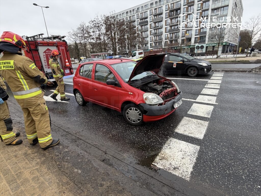 Czerwony samochód z otwartą maską na pasie drogowym, obok strażacy w mundurach przygotowujący się do interwencji.