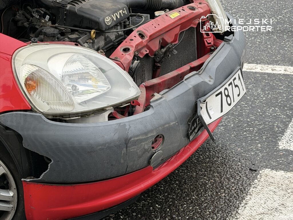 Czerwony samochód osobowy z uszkodzonym zderzakiem stojący na parkingu, widoczny fragment silnika.