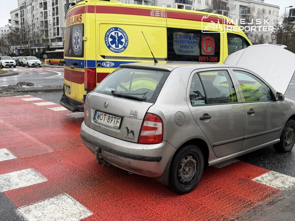 Na skrzyżowaniu stoi uszkodzony samochód osobowy z otwartą maską, obok niego zatrzymana karetka pogotowia.