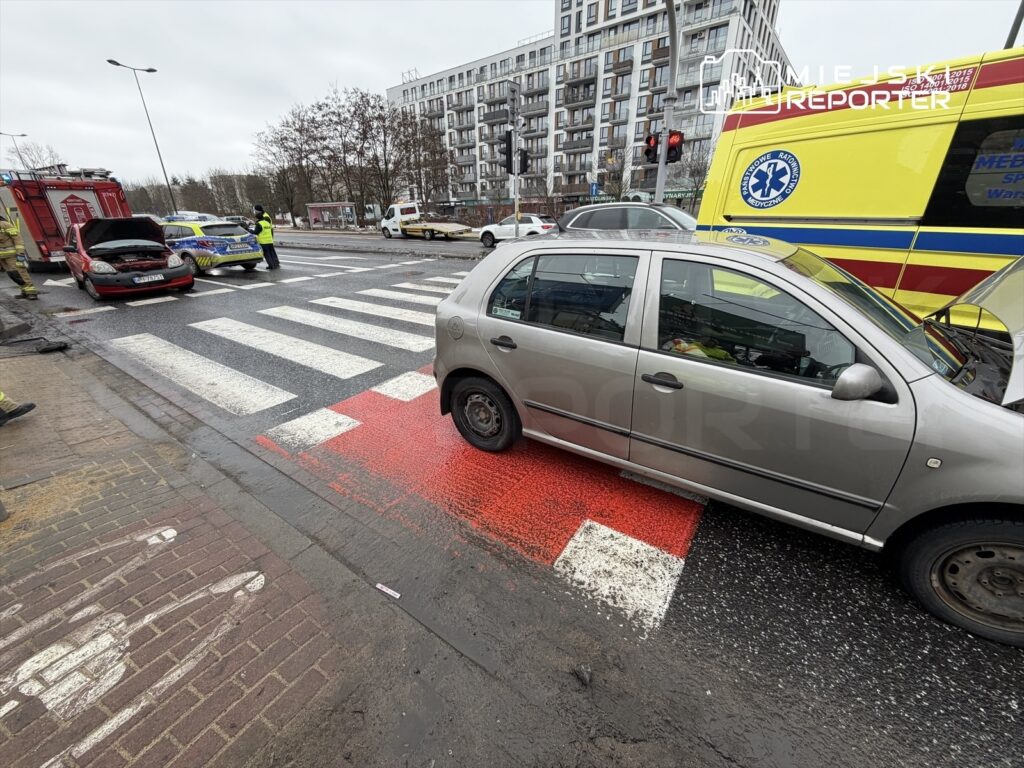 Na pasach dla pieszych stoi uszkodzony samochód osobowy, obok niego zespół ratunkowy oraz straż pożarna. W tle widać nowoczesne budynki.