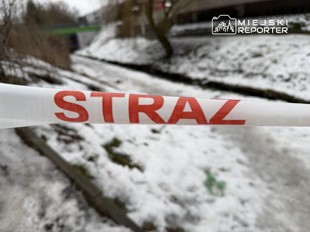 Taśma zabezpieczająca z napisem "STRAŻ" rozciągnięta wzdłuż zamarzniętego cieku wodnego w zimowym otoczeniu.
