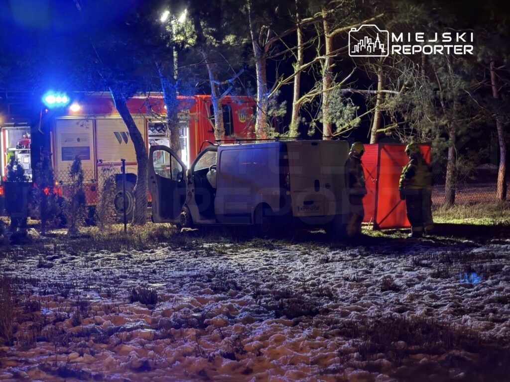 Strażacy w żółtych hełmach pracują obok białego vana w lesie, w tle widoczny wóz strażacki z włączonymi sygnałami.