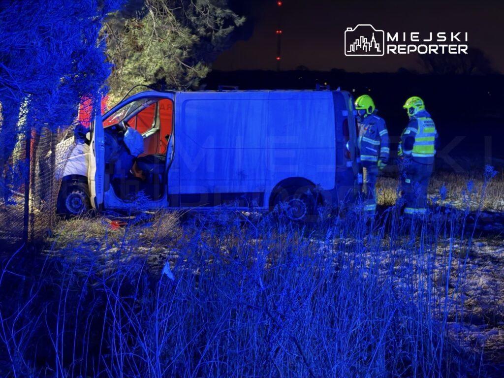 Dwa ratowników w odblaskowych kamizelkach pracują przy rozbitym białym vanie w ciemnym terenie, oświetlonym niebieskimi światłami.