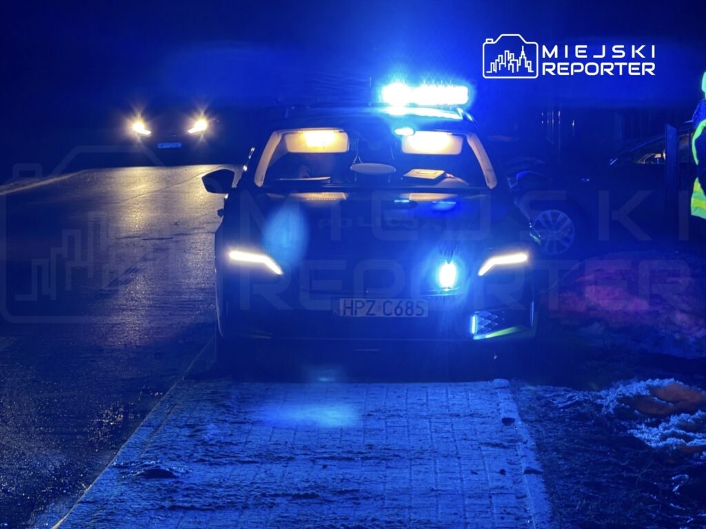 Radiowóz policyjny z włączonymi niebieskimi światłami stoi na oświetlonej ulicy, obok widać drugie auto.