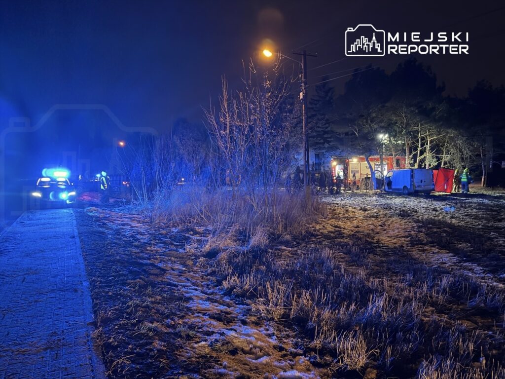 Radiowóz policyjny z włączonymi sygnałami świetlnymi stoi na poboczu, obok działa ekipa ratunkowa przy wozie strażackim.