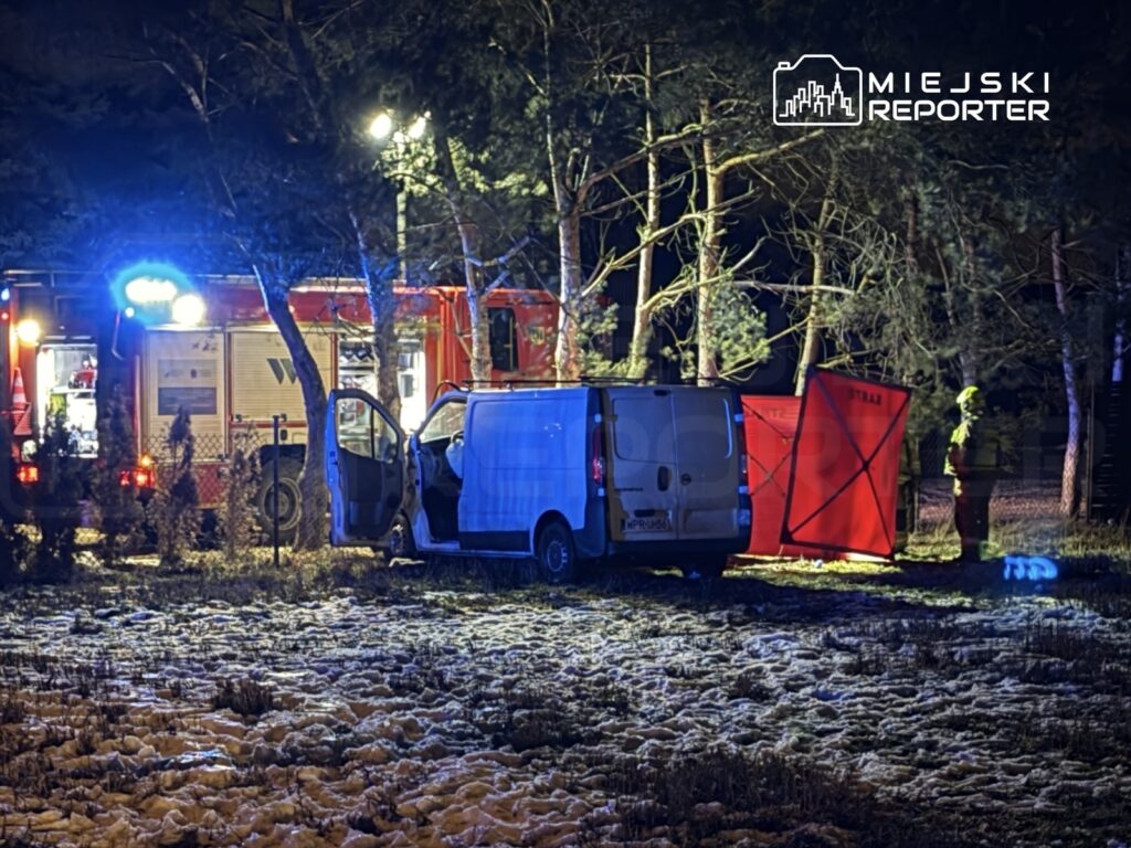 Ratownicy w żółtych kombinezonach pracują przy białym vanie, obok czerwonej osłony, w ciemnym lesie.