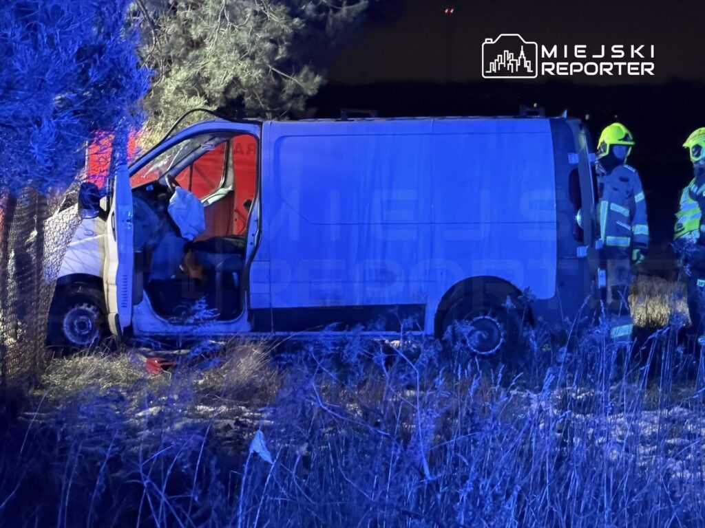 Biały dostawczy samochód zaparkowany w trawie, obok stoją strażacy w hełmach i mundurach, otwierają drzwi pojazdu.