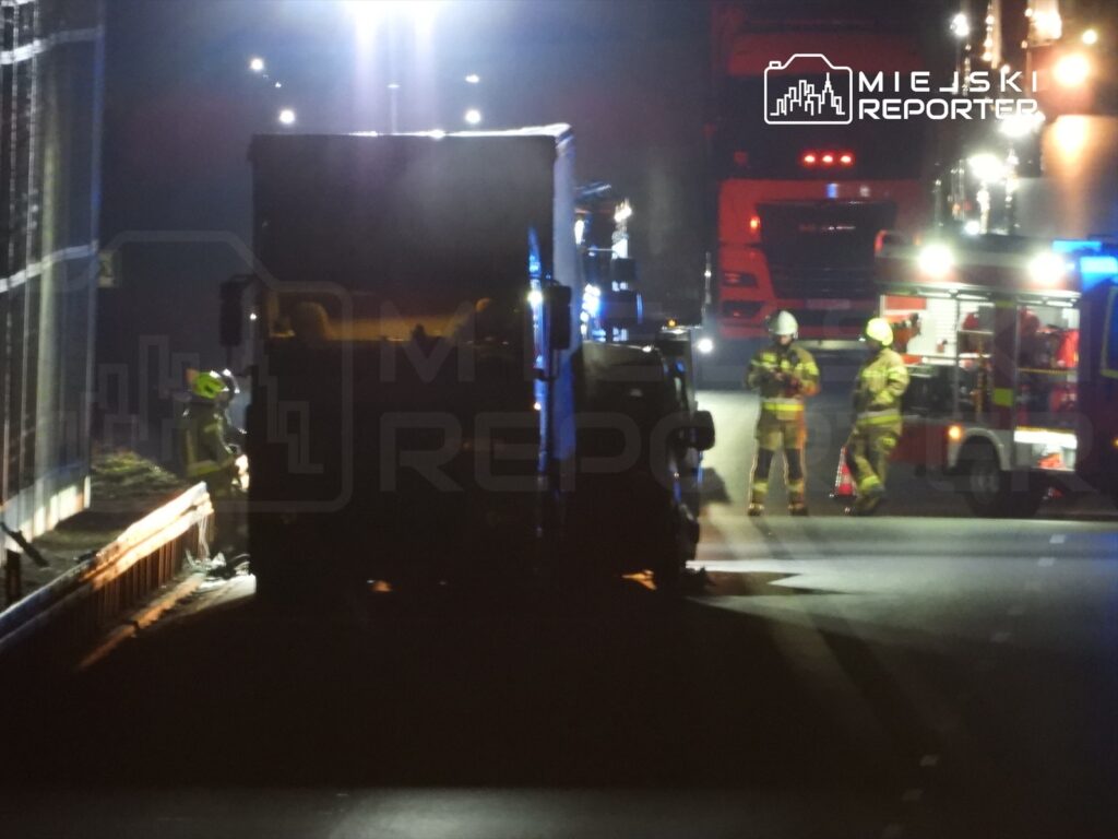 Strażacy w hełmach oceniają sytuację przy przewróconej ciężarówce na drodze, w tle widać inne pojazdy.