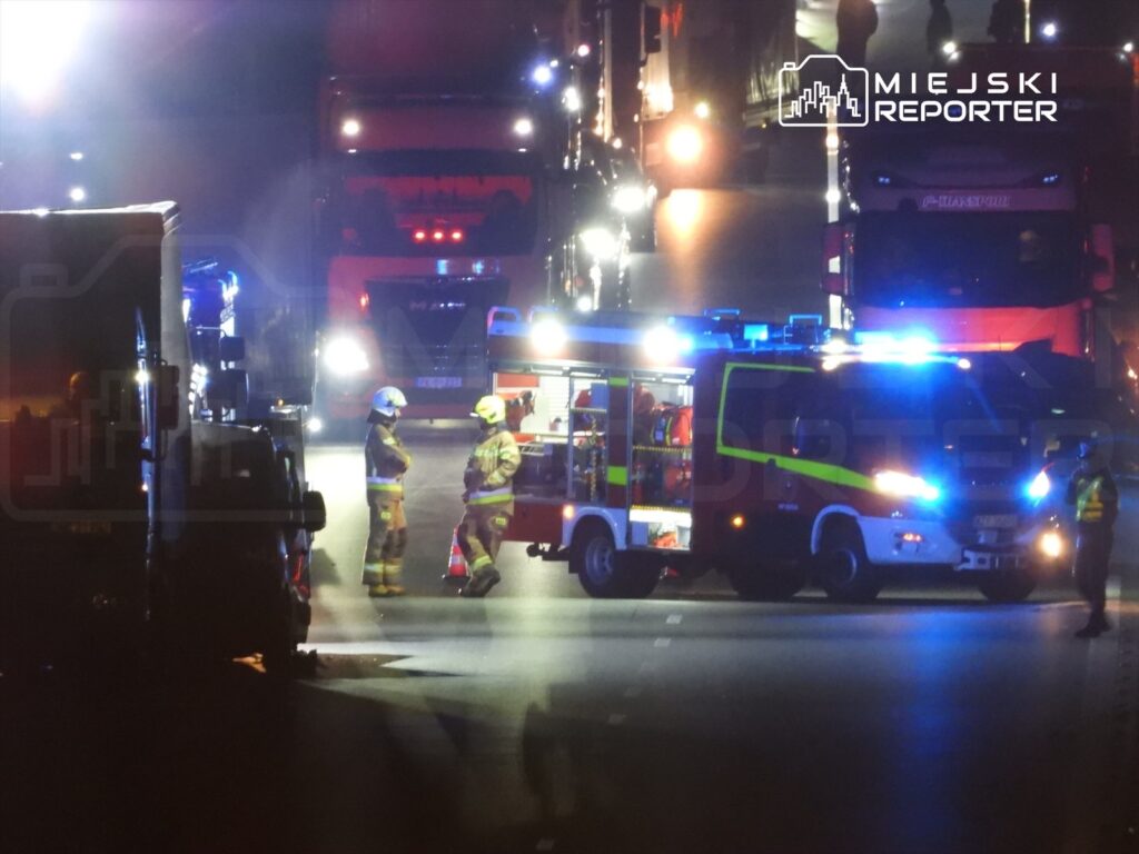 Strażacy rozmawiają obok wozu strażackiego na drodze, w tle ciężarówki z oświetleniem nocnym.