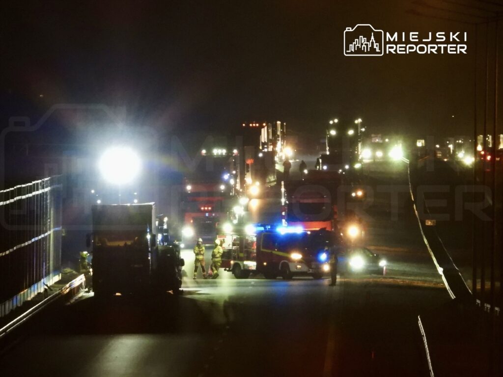 Grupa strażaków i ratowników pracuje przy oświetlonym wozie strażackim na drodze, w tle widoczne ciężarówki.