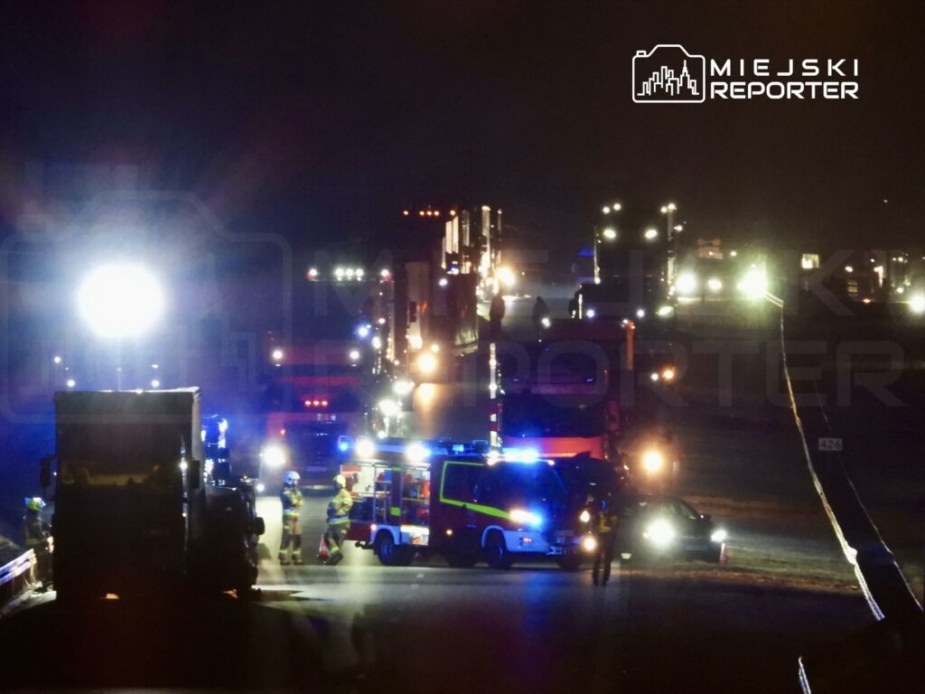 Na drodze nocą zgrupowane pojazdy ratunkowe oraz ciężarówki, w tle widoczne światła i strażacy interweniujący przy wypadku.