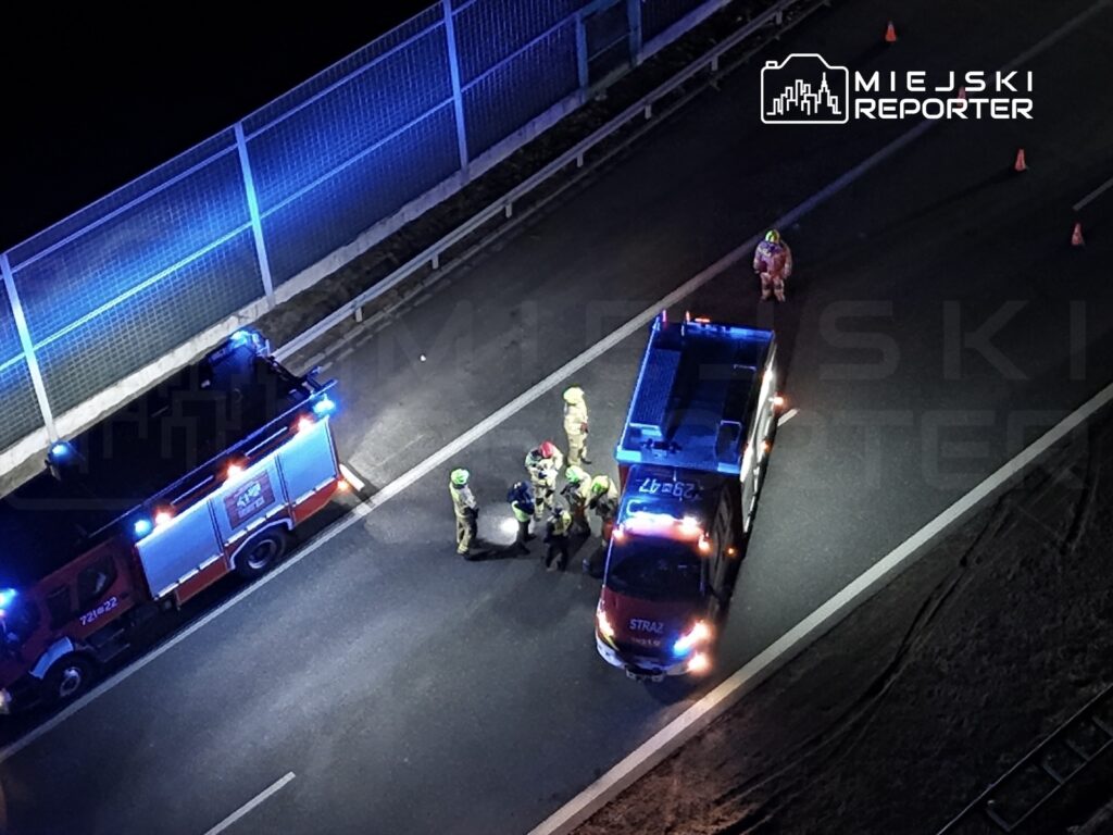 Grupa strażaków w kaskach zbiera się wokół pojazdu na drodze, obok czerwonego wozu strażackiego z włączonymi sygnałami.