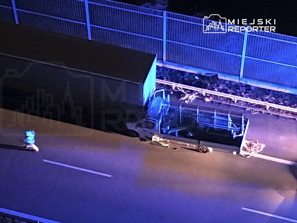 Mężczyzna w niebieskiej kurtce biegnie obok zatrzymanego ciężarowego pojazdu na oświetlonej drodze nocą.